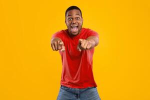 Hey, You. Emotional Black Guy Pointing Fingers At Camera With Both Hands And Shouting In Excitement Posing Standing Over Yellow Studio Background, Wearing Bright T-Shirt. You're Next Concept photo
