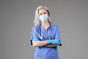 A mature lady doctor stands confidently, wearing blue scrubs, a medical mask, and gloves. She has long gray hair and is ready for a healthcare challenge in a professional setting. photo