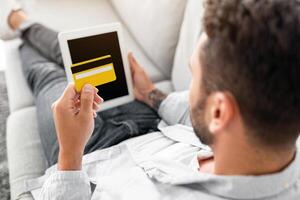 E-commerce concept. Unrecognizable man with credit card and digital tablet sitting on sofa at home, over shoulder shot. Young guy shopping online, using modern pad and holding card photo