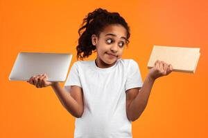 Portrait of doubtful puzzled black schoolgirl holding digital tablet or laptop and paper book, comparing and choosing between paper manual vs modern technology, isolated on yellow studio background photo