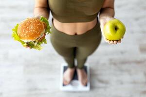 Above view of unrecognizable Indian woman holding apple and hamburger, standing on scales, choosing diet. Young lady selecting nutrition, making hard choice, closeup view photo