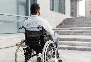 Back view of young black guy in wheelchair standing in front of stairs without ramp, having problem entering building, empty space. Impairment and accessible city environment concept photo