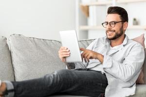 Technology concept. Smiling millennial man using digital tablet resting on couch at his modern apartment, free space. Happy guy browsing internet on pad, resting on sofa in living room photo