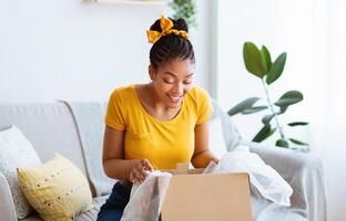 Satisfied Buyer. Portrait of curious cheerful black woman received package, unpacking cardboard box, looking inside, sitting on the sofa in living room at her apartment. Happy with online purchase photo