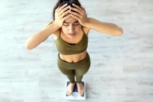 Unsuccessful slimming and dieting concept. Upset young Indian woman unhappy about her weight, standing on scales, holding head in disappointment indoors, above view photo