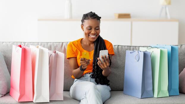 en línea compras y entrega. positivo negro mujer con crédito tarjeta y Teléfono móvil sentado en sofá con regalo bolsas, confirmando su Internet orden. milenario africano americano dama adquisitivo en web foto