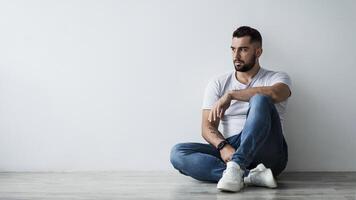 Lonely handsome man sad, stressed in self-isolation, suffering agoraphobia, depression. Serious millennial attractive muscular man sitting on floor, on gray wall background and looking at empty space photo