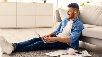 Home Office Concept. Smiling Jew Man In Wireless Earphones Sitting On Rug Floor Carpet, Working On Laptop Indoors In Living Room. Happy Guy In Kipa Using Computer. Distant Job, Internet And Technology photo
