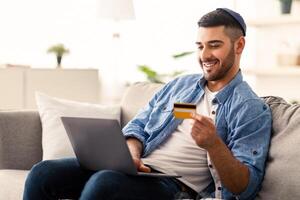 Making Financial Transaction. Smiling young jewish guy in kipah holding and showing debit credit card, sitting on couch with laptop computer at home, selective focus, blurred background photo
