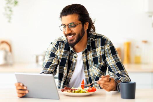 hermoso pelo largo, barbado árabe chico teniendo desayuno, comiendo Fresco verduras, salchichas, Bebiendo café y utilizando moderno digital tableta, acecho serie o videos en línea, cocina interior foto