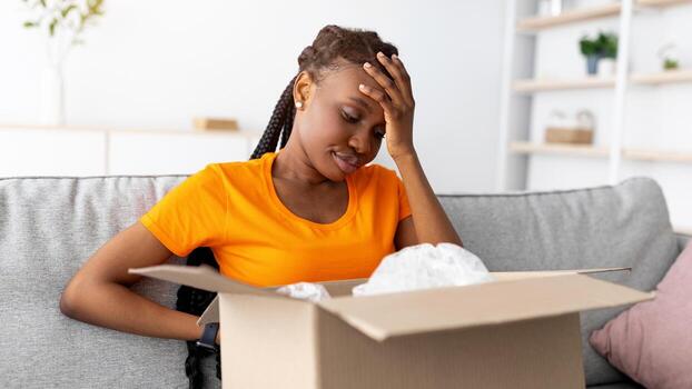 dañado paquete. decepcionado africano americano mujer desembalaje su entrega, mirando dentro incorrecto caja, infeliz porque de Envío error, conmovedor su frente en frustración. en línea compras foto