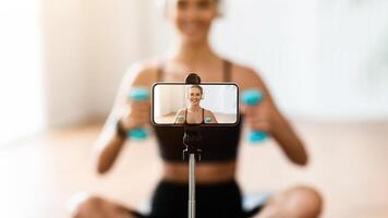 Fitness Blog. Happy woman in headphones exercising with dumbbells in front of smartphone on tripod at home, free copy space, selective focus on gadget screen. Fitness blogger recording tutorials photo