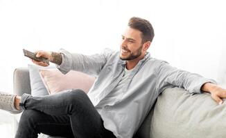 Stay home entertainments. Relaxed man with remote control watching TV on sofa in living room, free space. Millennial guy with television controller switching channels on couch indoors photo
