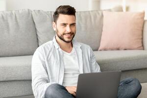 Remote job concept. Successful young freelancer man working on laptop computer, sitting on floor and leaning on sofa, working remotely from home, free space photo