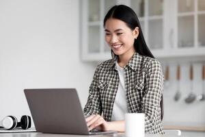 Positive asian lady freelancer sitting at kicthen, working online, using modern laptop, typing on keyboard, sending emails or chatting with clients. Young woman student doing homework, copy space photo