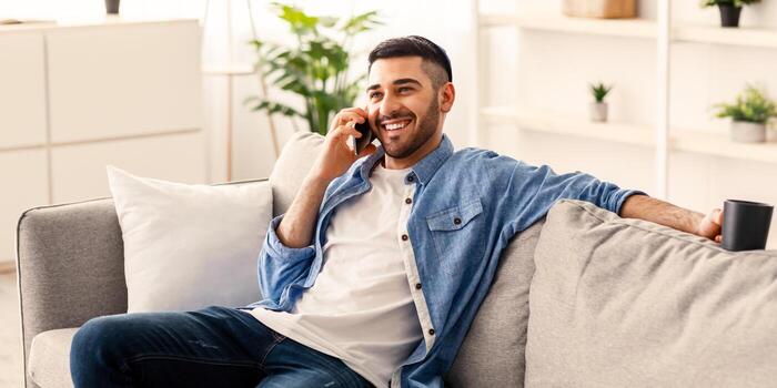 móvil comunicación concepto. retrato de sonriente joven judío chico en kipa hablando en Teléfono móvil, participación taza Bebiendo café, trabajando remotamente sentado en cómodo sofá con ordenador portátil a hogar oficina foto