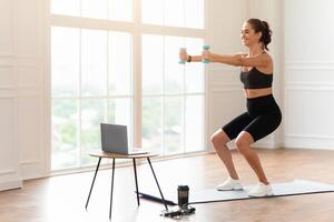 Positive fit lady exercising and squatting with two dumbbells in front of pc on coffee table at home, smiling fitness blogger recording for her followers having personal virtual training photo
