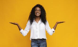 Make right choice. Positive african girl comparing variants, making scales with his empty hands, yellow studio background, free space photo