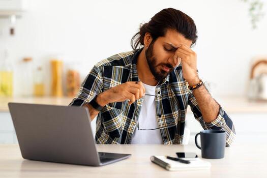 cansado Medio este joven hombre persona de libre dedicación teniendo dificultades a trabajo, sentado a cocina mesa con computadora portátil, teléfono inteligente, bloc, participación anteojos, frotamiento nariz puente, Copiar espacio. profesional agotamiento foto