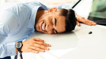 Happy young arab man hugging car of his dream, closing eyes and smiling, closeup photo, side view, copy space. Ecstatic middle eastern guy found his auto, visiting automobile salon photo