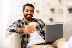 Positive long-haired, bearded indian guy freelancer sitting on sofa with laptop, typing on computer keyboard and using cellphone, working from home, looking at screen and smiling, copy space photo
