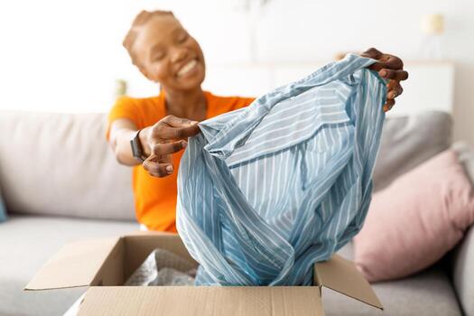 satisfecho comprador concepto. sonriente negro mujer desembalaje paquete después entrega, participación blusa, sentado en sofá a hogar. africano americano dama contento con en línea compra, selectivo atención foto