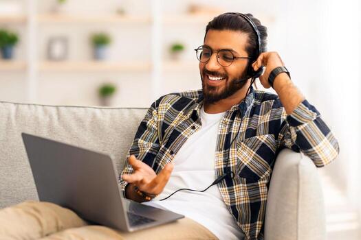 alegre pelo largo, barbado indio chico sentado en sofá, utilizando ordenador portátil y auriculares, teniendo conferencia con cliente o Novia desde hogar, sonriente y gesticulando a cuaderno pantalla, Copiar espacio foto