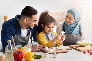 Portrait Of Cute Little Arab Girl Cooking With Parents In Kitchen, Happy Middle Eastern Family Of Three Preparing Healthy Tasty Food Together, Pretty Small Female Kid Holding Mushroom Slice In Hands photo