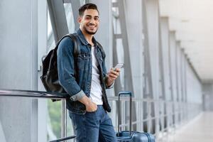 Portrait Of Cheerful Young Arab Guy Using Smartphone While Waiting At Airport, Happy Smiling Middle Eastern Man Browsing Internet On Cellphone While Standing With Luggage At Terminal, Copy Space photo