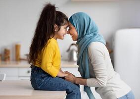 Mother's Day Concept. Adorable Little Girl Bonding With Her Muslim Mom In Hijab At Home, Happy Islamic Lady Holding Hands And Touching Foreheads With Cute Female Child, Side View With Copy Space photo
