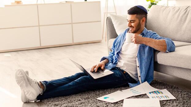 personas y tecnología concepto. retrato de sonriente joven judío hombre en kipa utilizando ordenador portátil participación taza y Bebiendo caliente café. masculino sentado en piso alfombra con papeleo, acecho , tomando descanso foto