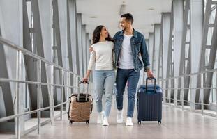 Trip Together. Happy Young Arab Spouses Walking With Luggage At Airport Terminal, Romantic Middle Eastern Couple Embracing And Smiling To Each Other While Going To Boarding Gate, Copy Space photo