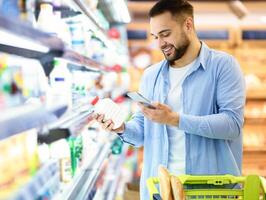 Smiling young man taking dairy products from shelf in the supermarket, holding bottle and smartphone, customer scanning bar code on product through mobile phone, walking with trolley cart photo
