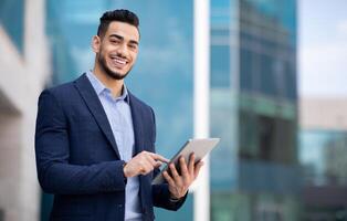 Good-looking arab guy in formal wear standing by office building, holding digital tablet and smiling, looking for new business opportunities online, using business application, copy space, panorama photo