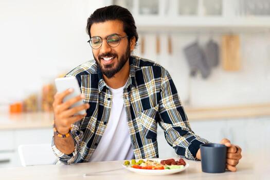 sonriente Medio este joven hombre comiendo sano comida y utilizando moderno móvil teléfono a cocina, Copiar espacio. contento árabe chico teniendo desayuno y chateando con su Novia en teléfono inteligente foto