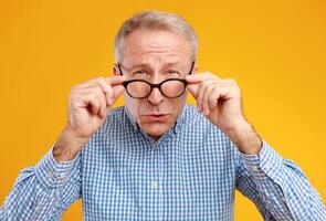 Poor Eyesight. Senior Man Can't See, Squinting Eyes Wearing Eyeglasses Having Problems With Vision, Looking At Camera, Yellow Orange Wall. Ophtalmic Issue In Older Age, Macular Degeneration Concept photo