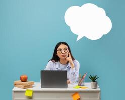 Pensive asian woman in glasses thinking with empty white speech bubble cloud with copy space, pondering questions, sitting at table isolated on blue studio background photo