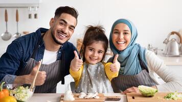 Cooking Together. Portrait Of Happy Muslim Family With Little Daughter Preparing Food In Kitchen, Adorable Small Girl Showing Thumbs Up At Camera, Enjoying Making Meals With Parents, Closeup photo