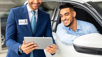 Rich middle-eastern businessman visiting car salon, sitting inside comfy white car and having conversation with cheerful sales assistant, looking at digital tablet screen in his hands, copy space photo