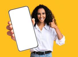 Cute pretty young middle-eastern woman showing modern mobile phone with empty screen and thumb up, closeup of cellphone with blank display, mockup, shot on yellow studio background, copy space photo