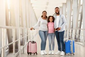 Happy Loving Family Of Three People Of Color Embracing In Airport Terminal Waiting For Flight, Cheerful Daughter Hugging Her Parents, Family Posing With Suitcases Holding Documents Looking At Camera photo