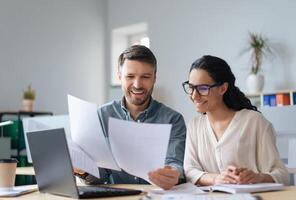 Cheerful male and female company workers looking through documents, collaborating, using laptop at modern office. Happy colleagues reading business papers, working on startup project together photo