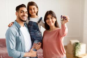 Positive family of three showing house key, standing in living room of new home, selective focus hand. Parents and daughter happy to move to bigger apartment, holding flat key, blurred background photo