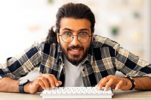Young middle-eastern man programmer sitting at worktable, using modern miniature pc keyboard, wearing glasses, closeup portrait. Happy arab guy developer working from home, enjoying his job photo