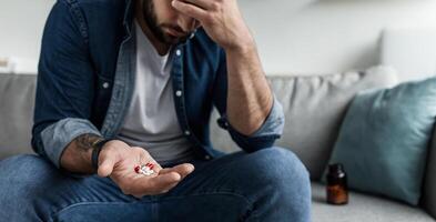 Commit suicide, health problems, depression and bad feeling. Pain, deep unhappy. Sad upset attractive young guy, hold many different pills in hand, hold his head in living room interior, copy space photo