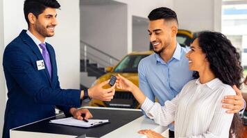 Happy middle-eastern couple buying car in huge showroom, taking automobile key from young handsome salesman, side view. cheerful arab man and woman purchasing beautiful auto, hugging and smiling photo