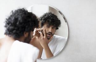 Problem skin. Concerned indian man popping pimple on cheek while standing near mirror in bathroom. Unhappy guy inspecting face, suffering acne, focus on reflection photo