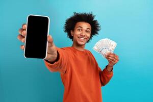A young African American man with curly hair smiles widely while holding a smartphone in one hand and cash in the other. He stands in front of a bright blue background, exuding joy and confidence. photo