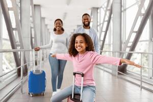 Domestic And International Flights Resumption. Portrait of black girl having fun, sitting on baggage and spreading arms, pretending air plane flying, playing while parent walking in blurred background photo