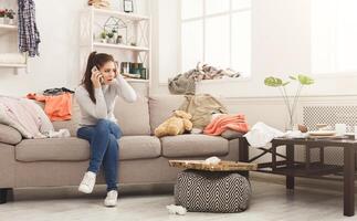 Desperate helpless woman sitting on sofa in messy living room. and talking on mobile, surrounded by many stack of clothes. Disorder and mess at home, copy space photo